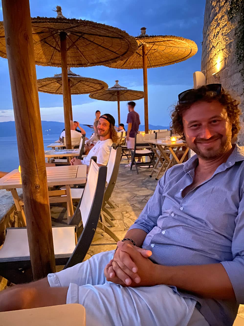 Ein Mann lächelt in die Kamera und sitzt in der Dämmerung in einem Restaurant am Meer. Die Kulisse besteht aus Holztischen, Strohschirmen und einem malerischen Blick auf das Meer mit einer Segelyacht in der Ferne. Im Hintergrund sind andere Gäste zu sehen.