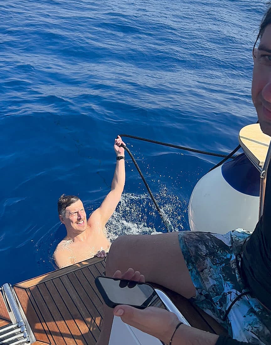 Ein Mann schwimmt im tiefblauen Ozeanwasser und hält sich dabei an einem Seil fest, das an einem Boot befestigt ist. Im Vordergrund ist eine weitere Person zu sehen, die auf dem Boot sitzt und ein Smartphone hält. Die Szene erinnert an einen gemütlichen Segeltörn während eines aufregenden Segelurlaubs auf dem Meer.