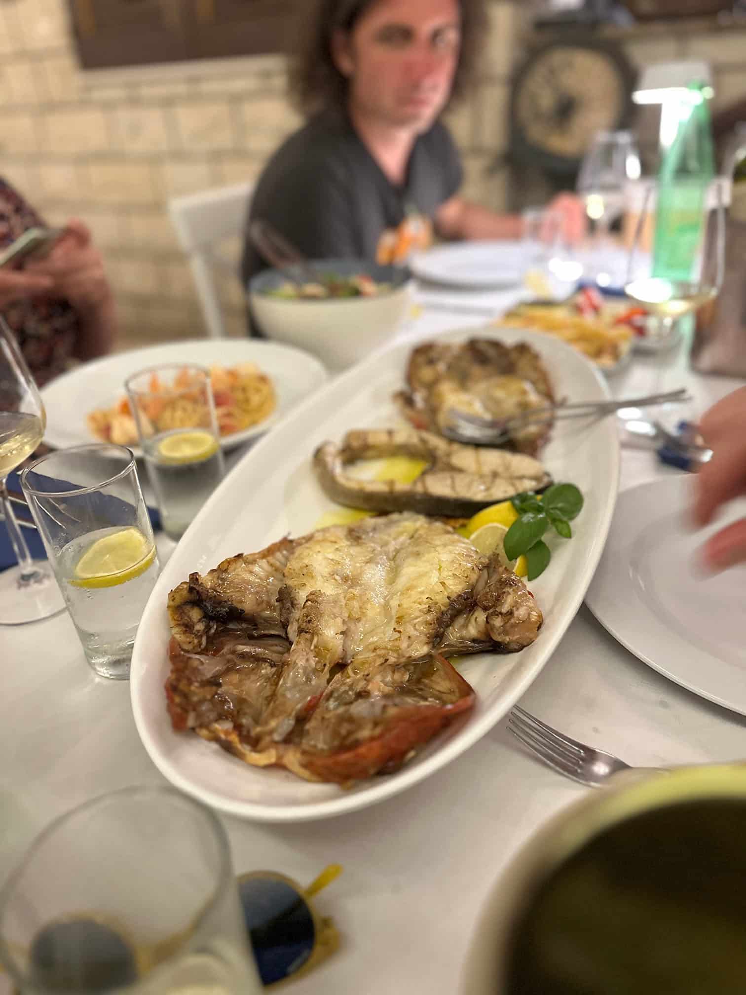 Ein Esstisch auf einer Segelyacht ist mit einer großen Platte mit gegrilltem Fisch und Zitronenscheiben gedeckt. Verschiedene Gerichte und Getränke stehen um die Platte herum. Im Hintergrund sitzt eine Person mit langen Haaren, und andere Gäste sind teilweise sichtbar. Die Kulisse scheint eine zwanglose Mahlzeit während eines ruhigen Segeltörns zu sein.