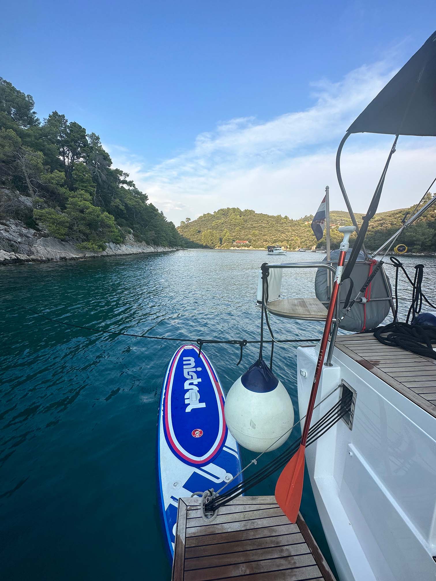 Ein Paddleboard mit der Aufschrift „Midnight“ liegt an der Seite einer eleganten Segelyacht. Ein Paddel und eine weiße Boje sind ebenfalls angebracht. Das Boot ist von einem ruhigen, klaren Gewässer umgeben, das von üppigen, grünen Bäumen unter einem bewölkten blauen Himmel gesäumt ist.