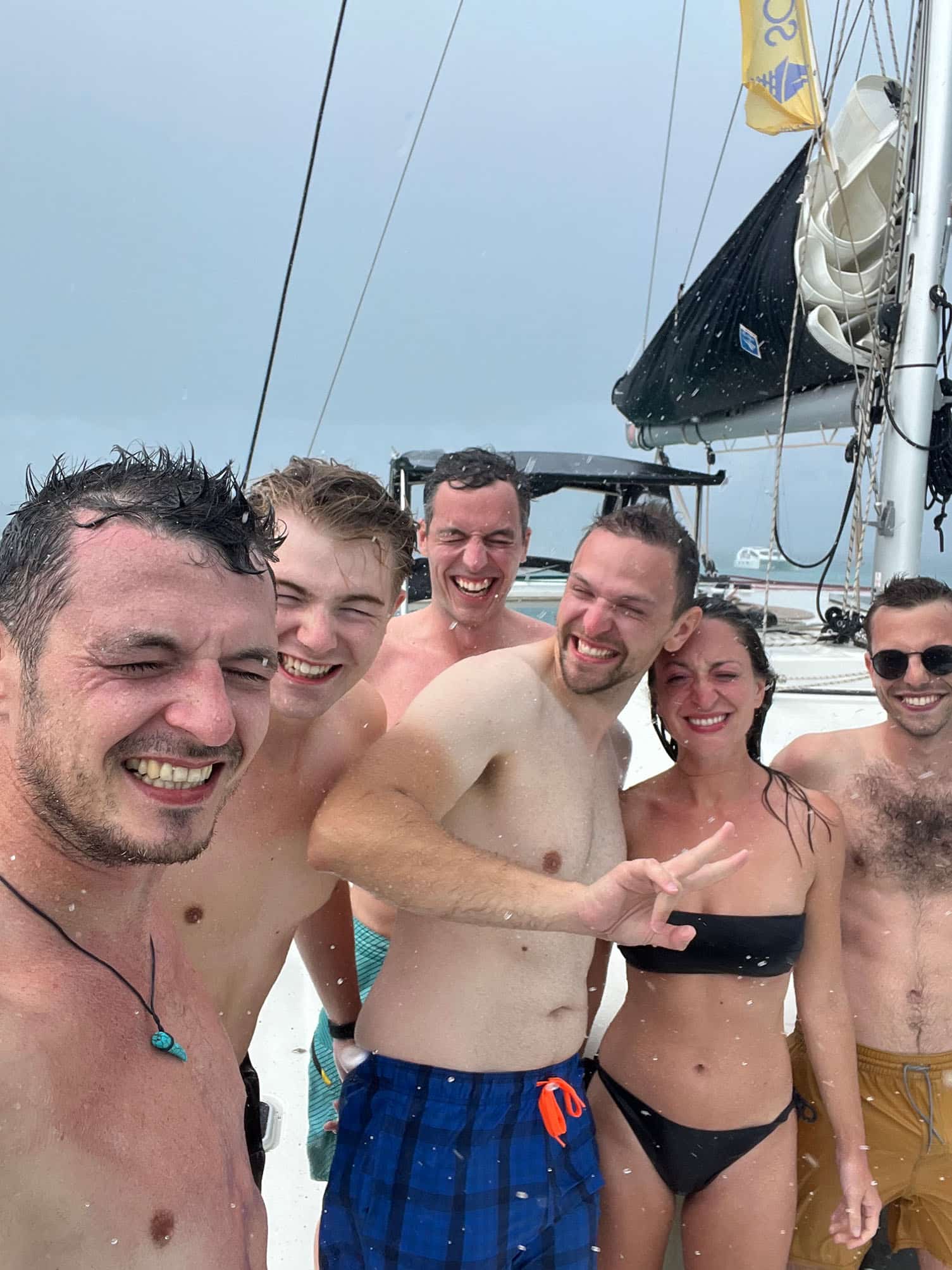Sechs Menschen lächeln während eines Regenschauers auf einer Segelyacht, tragen Badebekleidung und genießen das nasse Wetter.