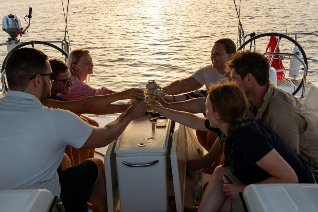 Sechs Personen sitzen um den Tisch auf dem Deck einer Segelyacht und stoßen an, während im Hintergrund die Sonne über dem Wasser untergeht. Sie scheinen einen entspannten und glücklichen Moment zusammen auf dieser unvergesslichen Segelreise zu genießen.