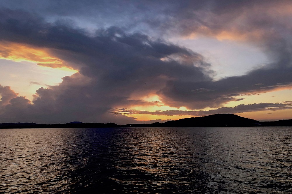 Sonnenuntergang über einem ruhigen Meer mit dramatischen Wolken in Orange-, Gelb- und Lilatönen, die den Horizont säumen, und Hügelsilhouetten im Hintergrund, beobachtet von einer Segelyacht.