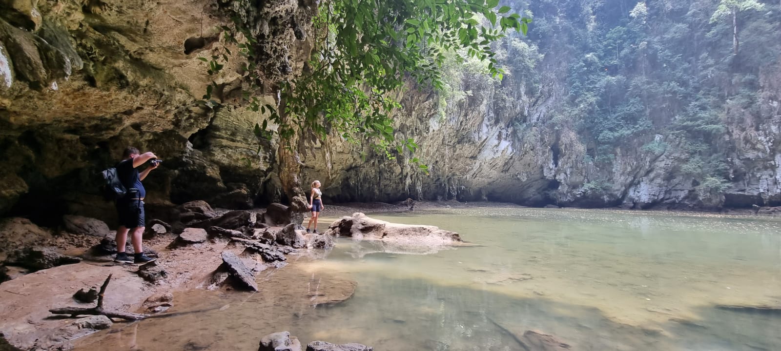 Eine Person macht Fotos in einer üppigen Höhle mit einem ruhigen Gewässer, felsigem Gelände und hängendem Laub, während andere auf ihrem Segelurlaub die Umgebung erkunden.