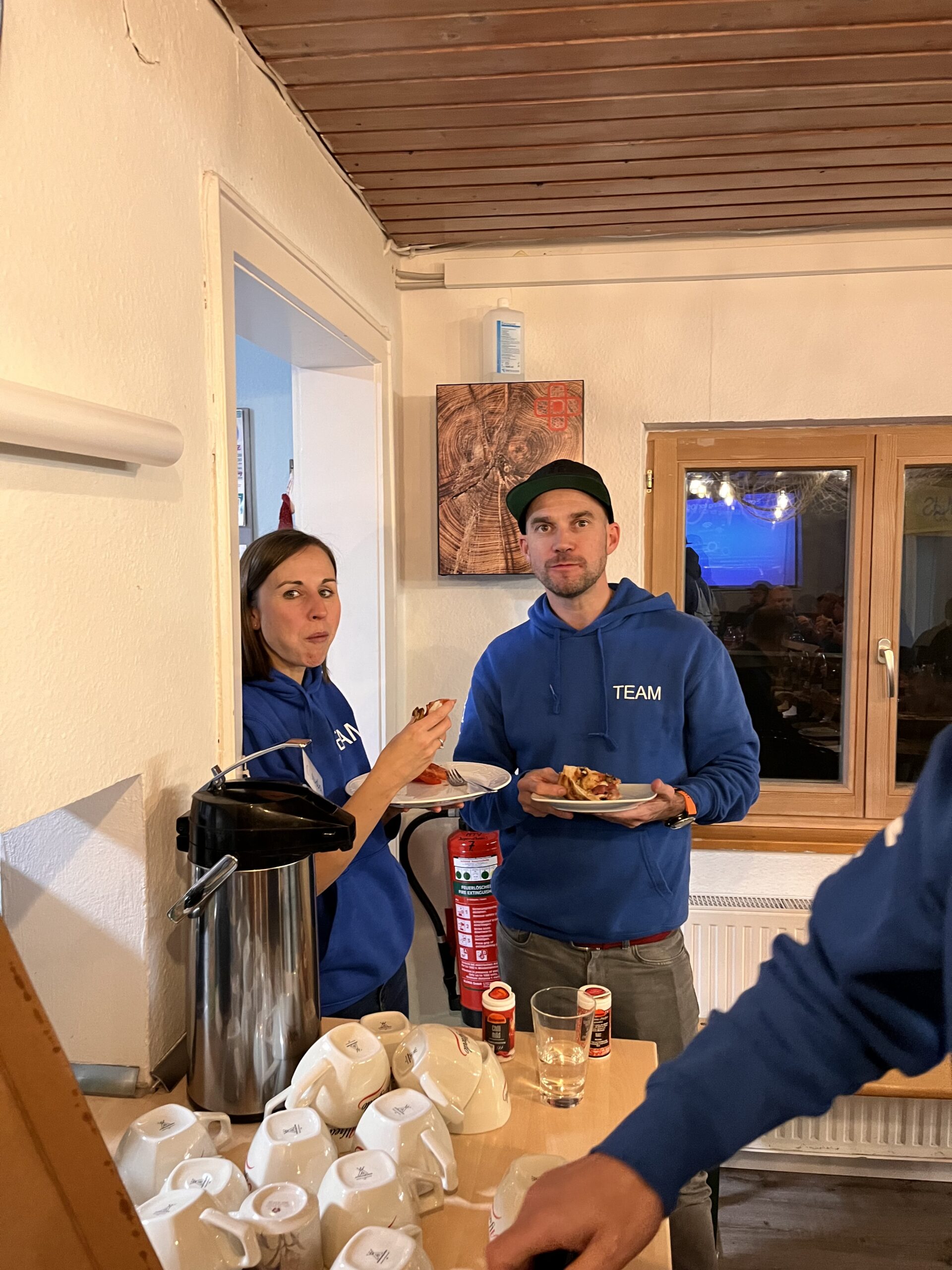 Zwei Personen in blauen „TEAM“-Hoodies stehen und essen neben einer Kaffeestation in einem Raum, der an eine gemütliche Segelyacht-Küche erinnert. Eine Person hält einen Teller und blickt in die Kamera, während die andere sich zu unterhalten scheint. Auf der Theke vor ihnen stehen Becherdeckel und ein Kaffeeautomat.