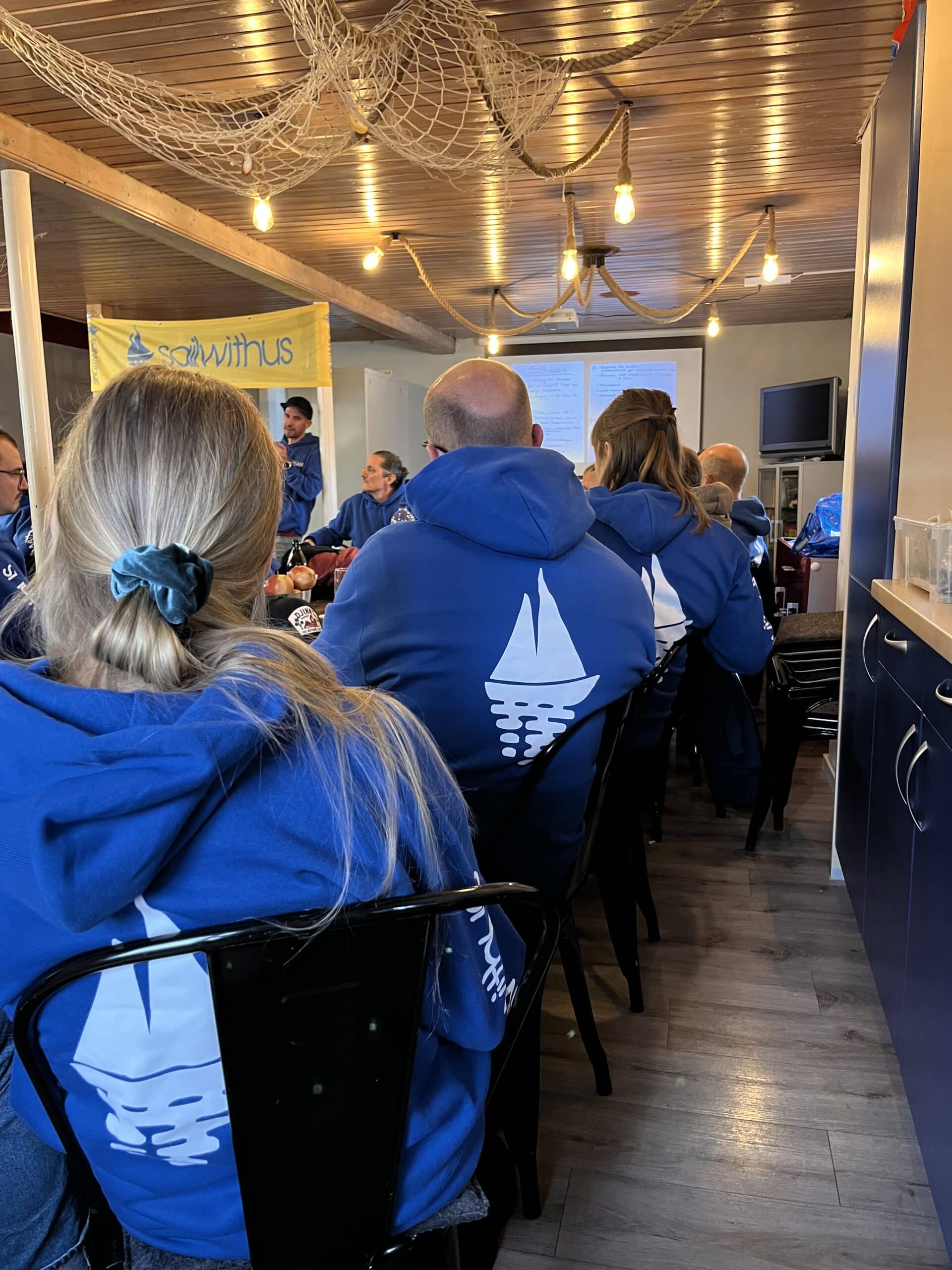 Eine Gruppe von Menschen in passenden blauen Kapuzenpullovern mit einem Segelboot-Logo sitzt in einem Raum mit Holzdecke und Beleuchtung und hört sich eine Präsentation an. Auf dem Bildschirm vorn werden Folien über ihre bevorstehende Segelreise angezeigt. Das lockere nautische Thema des Raums verstärkt die maritime Atmosphäre.