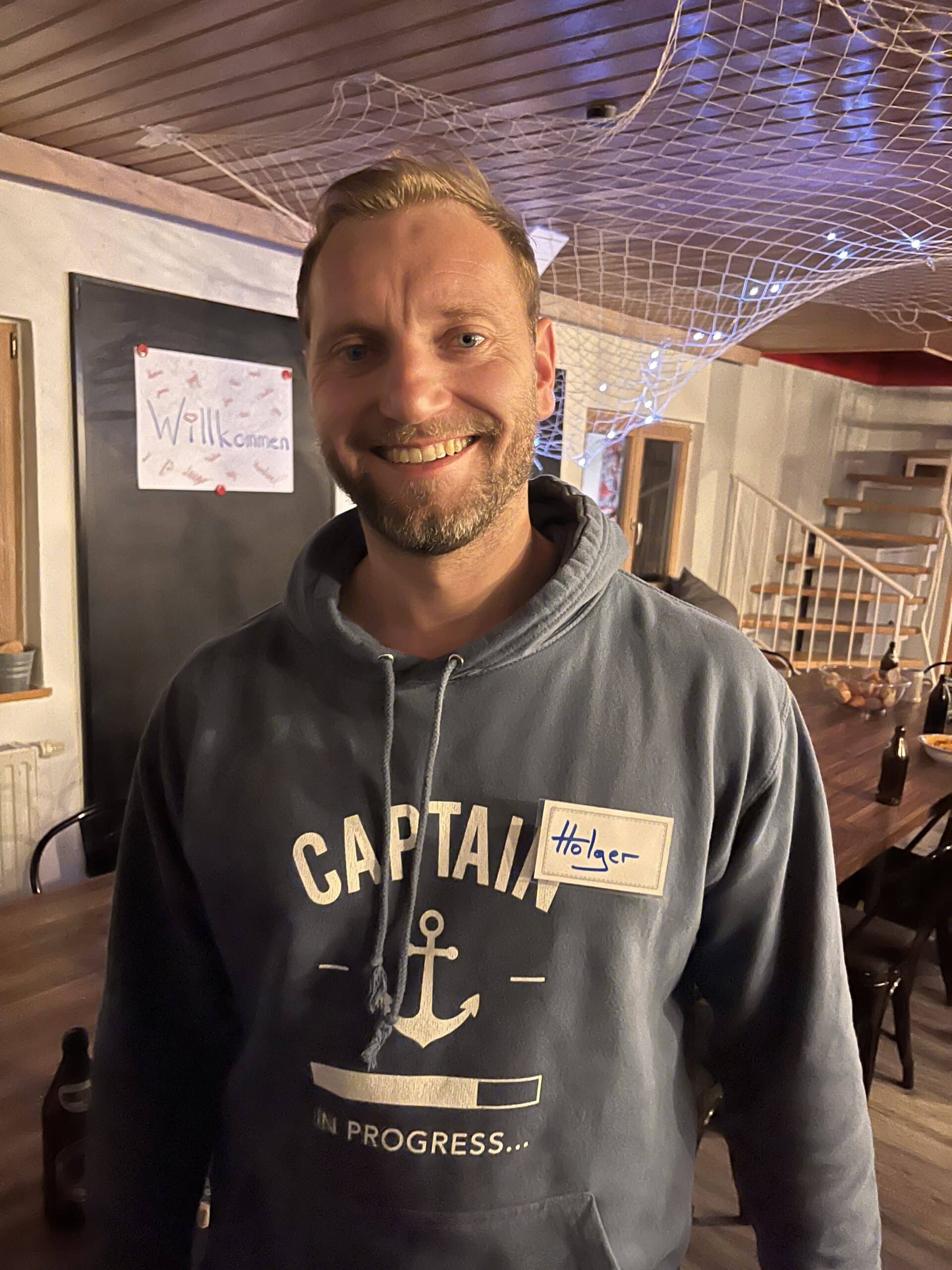 Ein lächelnder Mann mit einem Kapuzenpullover mit der Aufschrift „CAPTAIN in progress...“ und einem Ankermotiv steht in einem Raum, in dem Netze von der Decke hängen und verschiedene Katamaran-Modelle ausgestellt sind. Er trägt ein Namensschild mit der Aufschrift „Holger“.