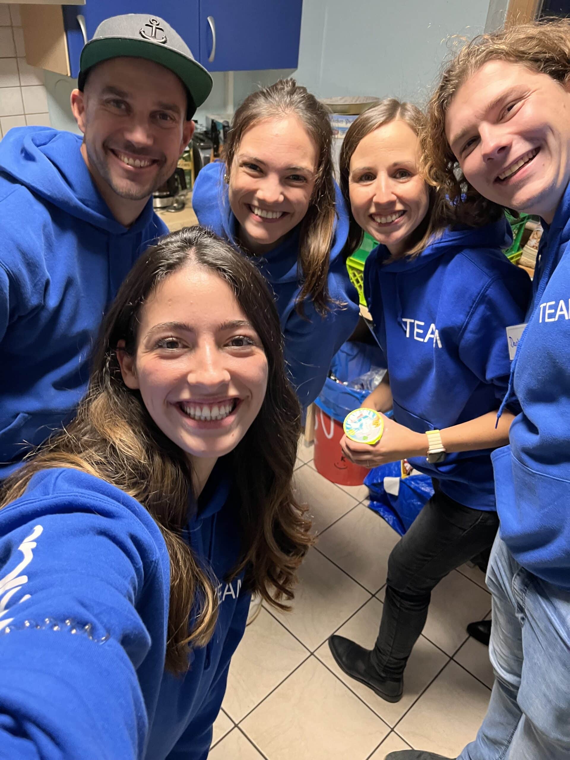 Fünf Menschen in blauen TEAM-Kapuzenpullis lächeln für ein Selfie in einer Küchenumgebung und verströmen eine fröhliche und gesellige Atmosphäre auf einer Segelyacht.