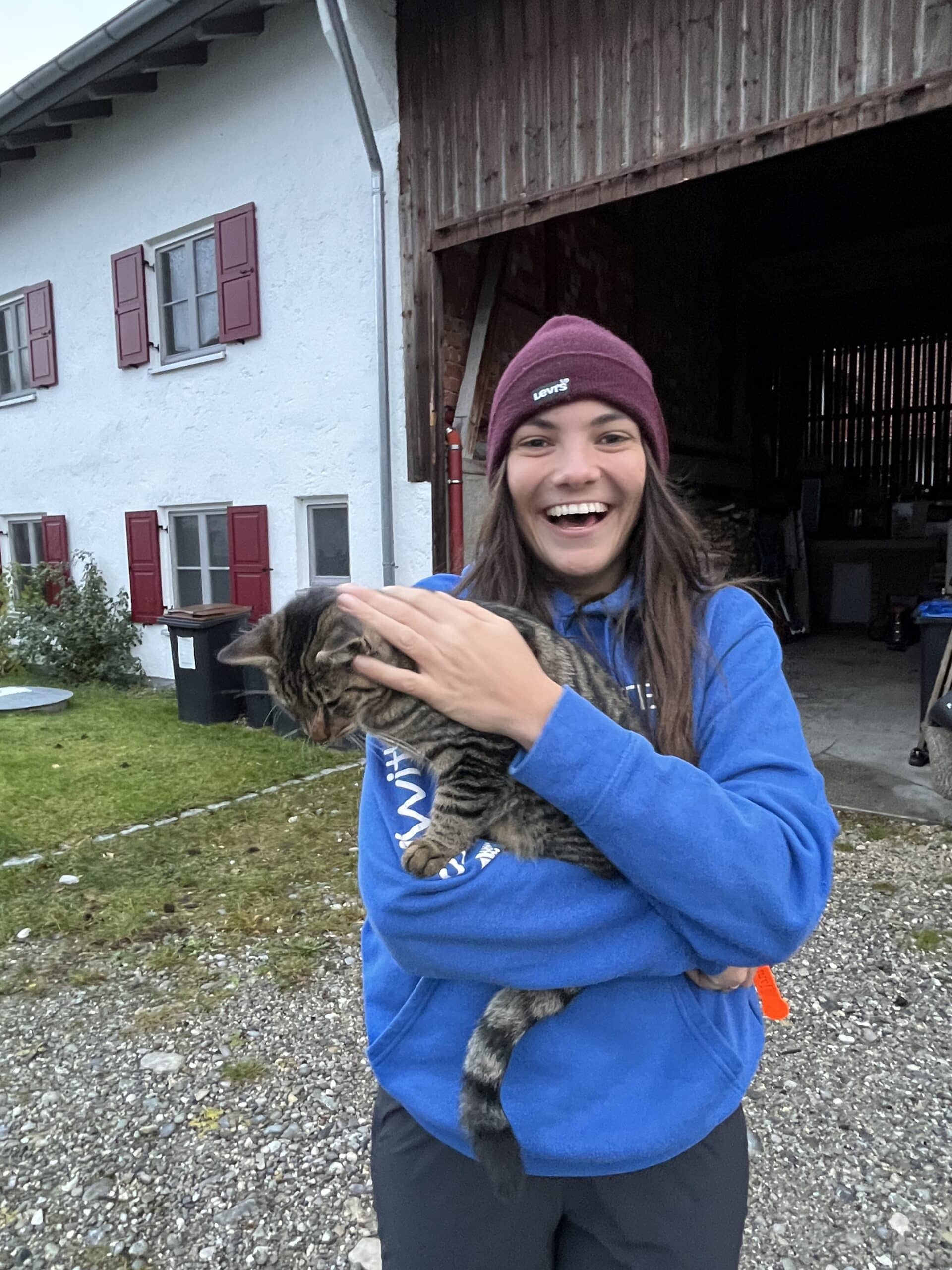 Eine fröhliche junge Frau mit blauem Kapuzenpullover und kastanienbrauner Beaniemütze hält eine zappelnde getigerte Katze vor einer Scheune mit roten Fensterläden an ihrem Segeltörn.