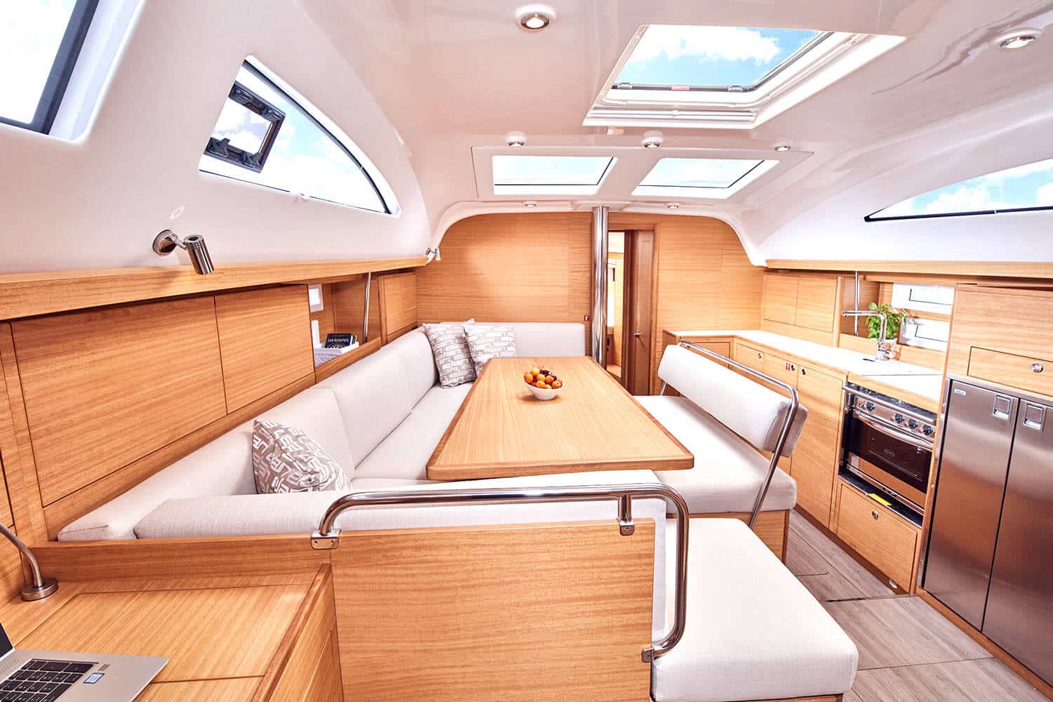 Innenansicht einer modernen Segelyacht mit einem gemütlichen Essbereich mit Holztisch, gepolsterten Bänken, reichlich natürlichem Licht durch Oberlichter und einem ausgestatteten Küchenbereich im Hintergrund.