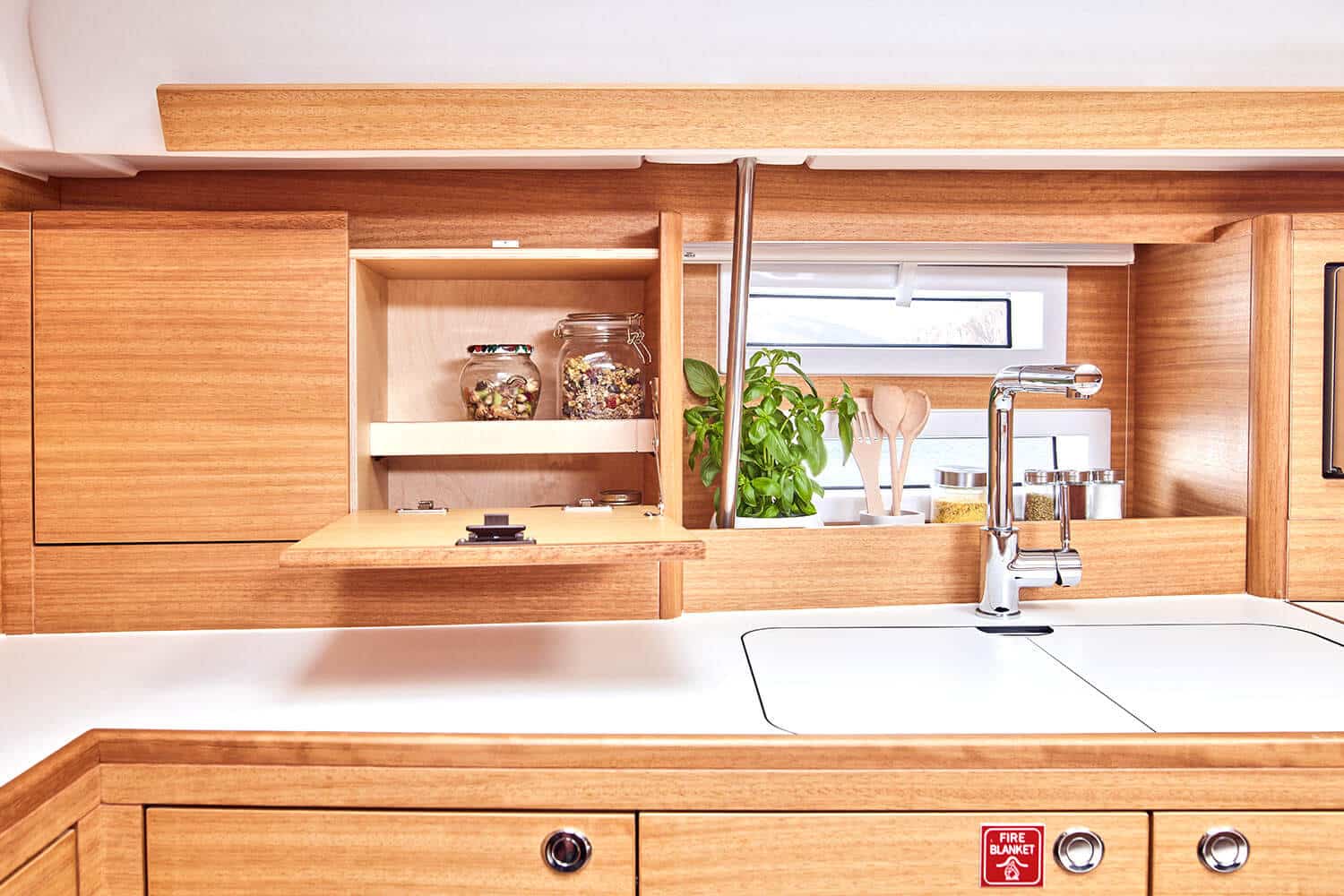 Ein sauberer Küchenbereich aus Holz im Inneren eines Katamarans. Ein Schrank mit einem ausziehbaren Regal stellt Gläser mit Zutaten zur Schau. Auf einer Arbeitsplatte stehen ein moderner Wasserhahn und eine Spüle, neben einem Fenster eine kleine Pflanze und Küchenutensilien. Darunter ist ein Schild mit der Aufschrift „FEUERLÖSCHER“ zu sehen.