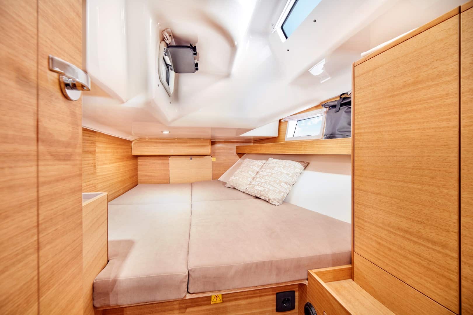 Eine gemütliche Bootskabine mit Holzwänden und eingebautem Stauraum erwartet Sie für Ihren Segelurlaub. Das Bett mit hellbrauner Bettwäsche hat zwei beige Kissen. Kleine Fenster lassen Tageslicht herein und eine Luke in der Decke ist zu sehen. Die Kabine verfügt über warmes Licht und ein klares, minimalistisches Design.