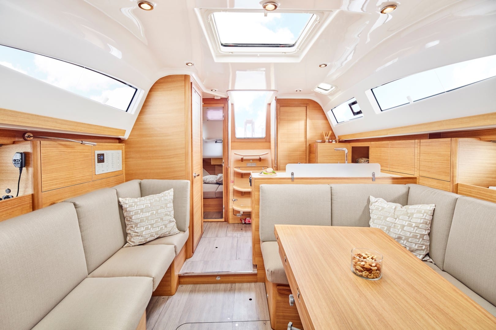 Innenraum eines modernen Segelboots mit heller Holzverkleidung und beiger Polsterung. Der Raum umfasst eingebaute Sitzgelegenheiten mit gemusterten Kissen, einen Holztisch mit einer Schüssel Snacks, Fenster oben und einen Blick in eine kleine, helle Schlafkabine – perfekt für Ihren nächsten Segelurlaub.