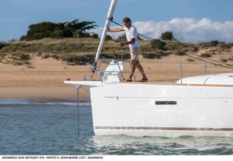 Eine Person in weißem Hemd und Shorts lichtet den Anker am Bug einer weißen Segelyacht Jeanneau Sun Odyssey 419. Das Boot liegt im seichten Wasser in der Nähe eines Sandstrands mit grasbewachsenen Dünen im Hintergrund und sorgt für einen idyllischen Segelurlaub unter einem klaren, sonnigen Himmel.
