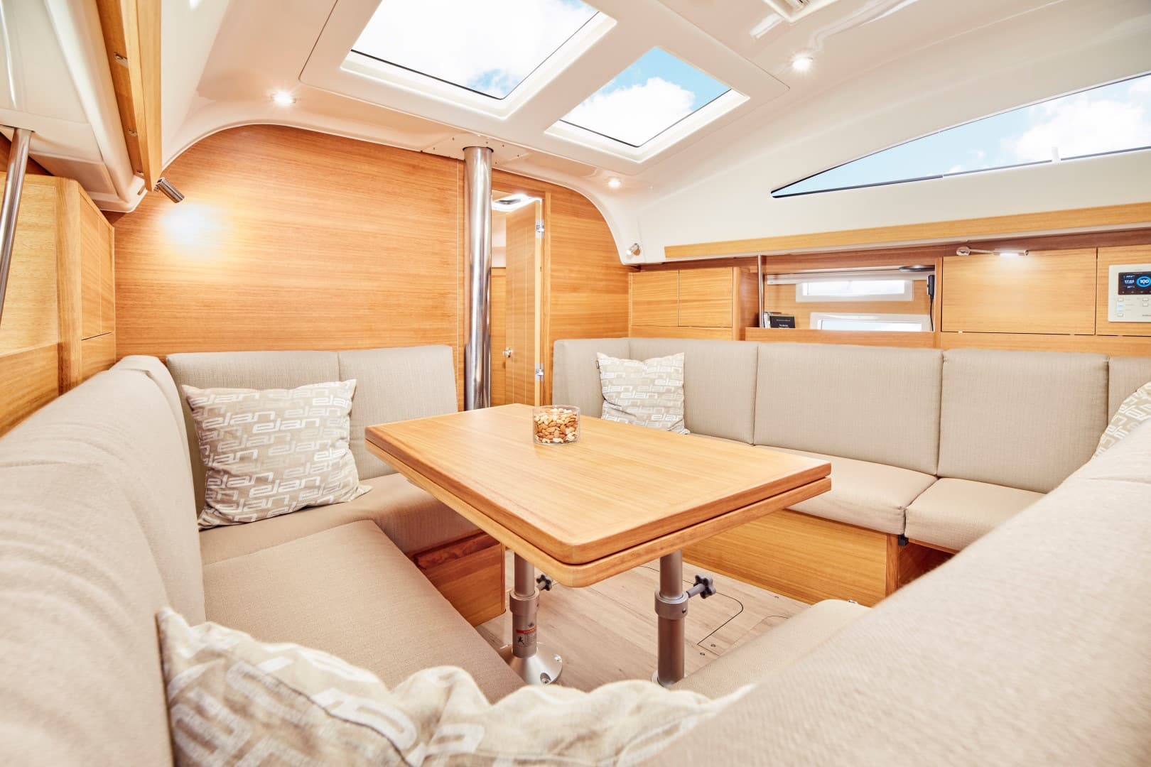 Innenansicht einer modernen Segelyacht mit gemütlichem Essbereich mit Holztisch, beige gepolsterten Sitzgelegenheiten und einer hellen Kabine mit Holzakzenten und großen Fenstern.