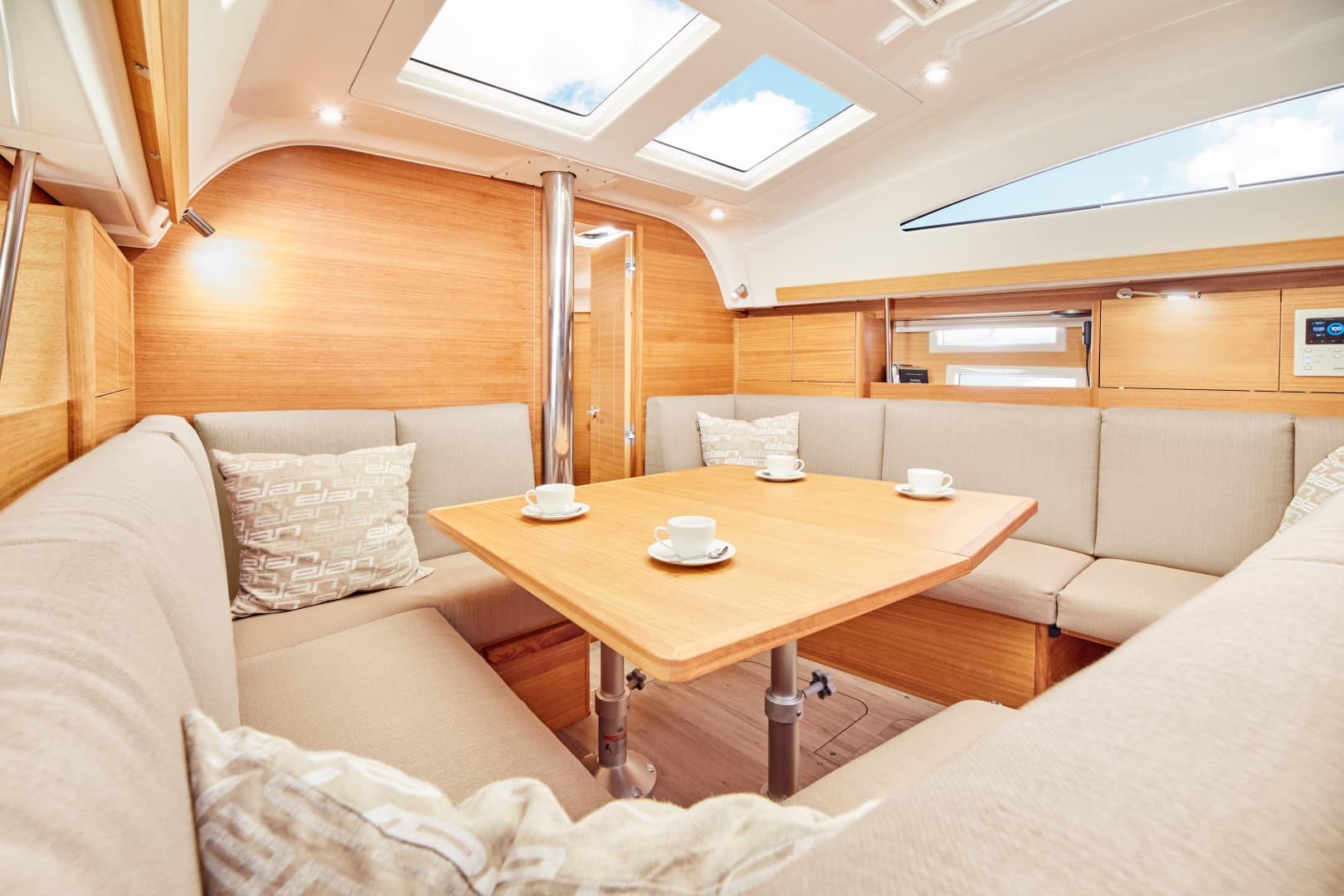 Innenansicht einer luxuriösen Segelyachtkabine mit einem Holztisch mit zwei Kaffeetassen, umgeben von plüschigen beigen Sofas, Holzvertäfelung und reichlich natürlichem Licht durch die Luken oben.