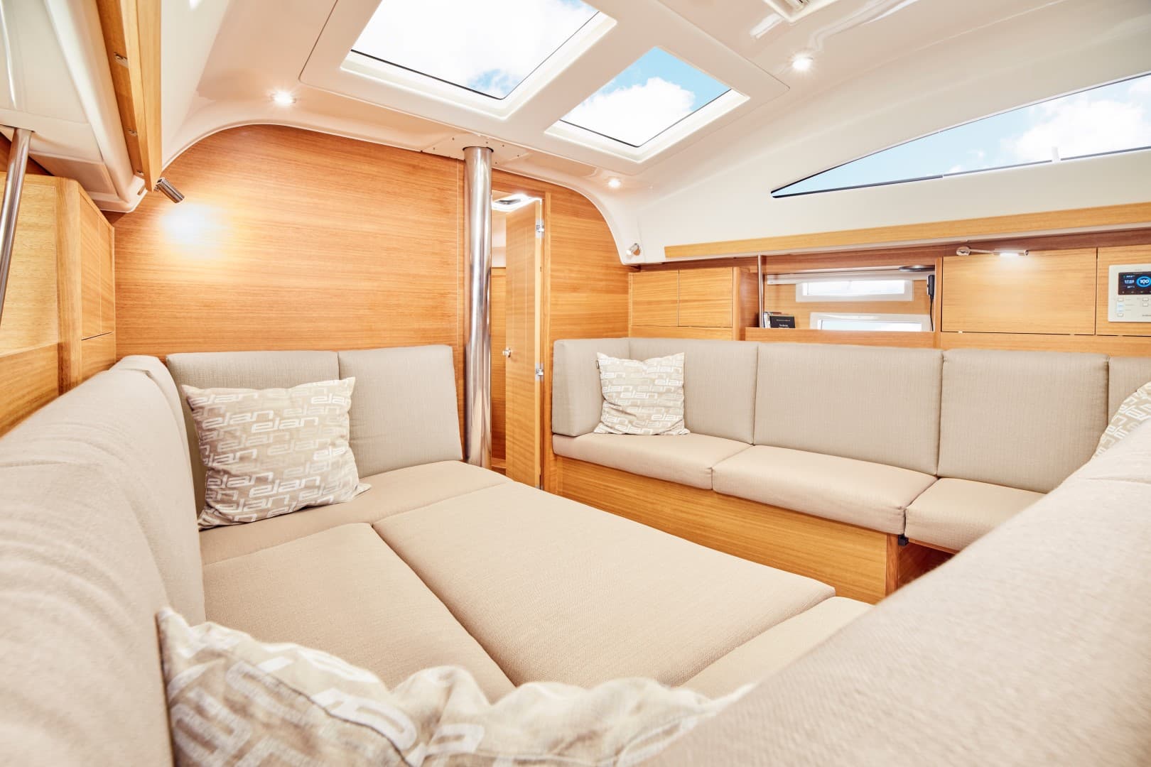 Das Bild zeigt ein modernes Bootsinneres mit geräumiger, heller Holzverkleidung. Es ist perfekt für einen Segelurlaub und verfügt über beige gepolsterte Sitze in L-Form mit dekorativen Kissen. Große Fenster und Oberlichter lassen natürliches Licht in den gemütlichen Raum, ideal für Ihre bevorstehende Segelreise.