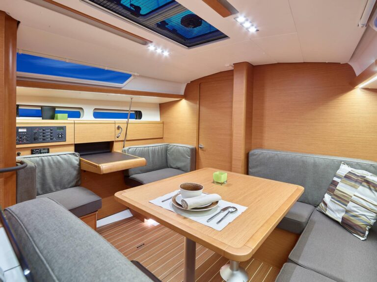 Innenansicht einer modernen Segelyachtkabine mit einem gemütlichen Essbereich mit einem Holztisch für zwei Personen, umgeben von gepolsterten Sitzbänken und sanfter Deckenbeleuchtung.