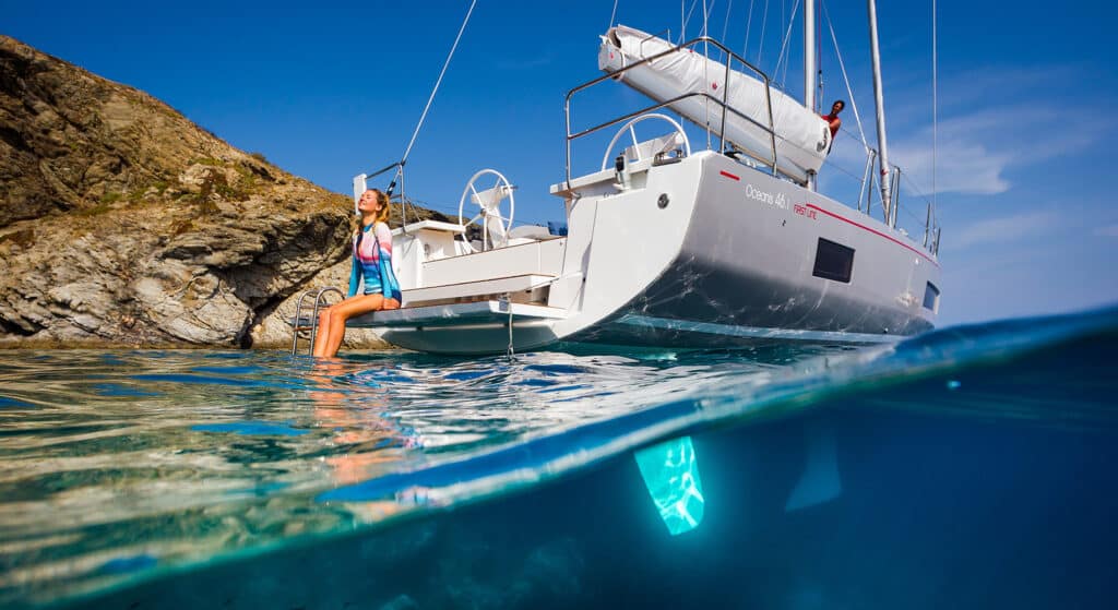Eine Frau steht auf dem Heck eines Katamarans in der Nähe von Felsklippen. Die eine Hälfte des Bildes zeigt die Unterwasseransicht und die andere Hälfte das sonnenbeschienene Boot und den klaren blauen Himmel.