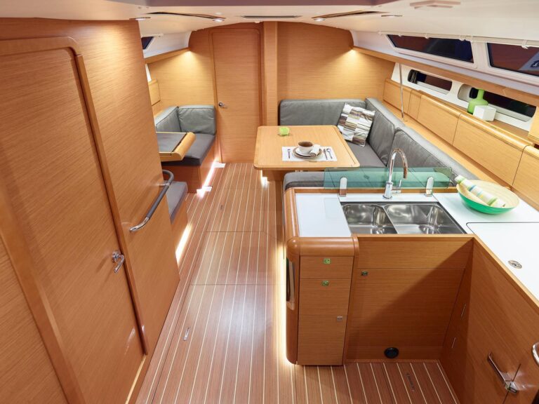 Innenansicht einer modernen Segelyachtkabine mit Holzboden, gemütlichem Essbereich, Küche mit Edelstahlspüle und großen Lagerschränken unter warmer Beleuchtung.
