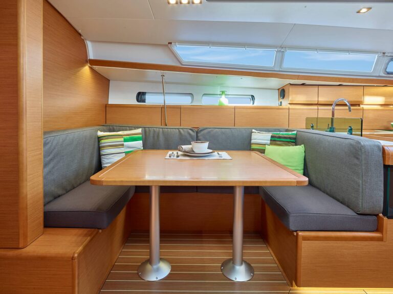 Innenansicht einer Segelyachtkabine mit Holzoberfläche, einem gemütlichen Essbereich mit einem braunen Tisch, grauen und grünen Kissen, einer Kaffeetasse und natürlichem Licht, das durch große Fenster fällt.