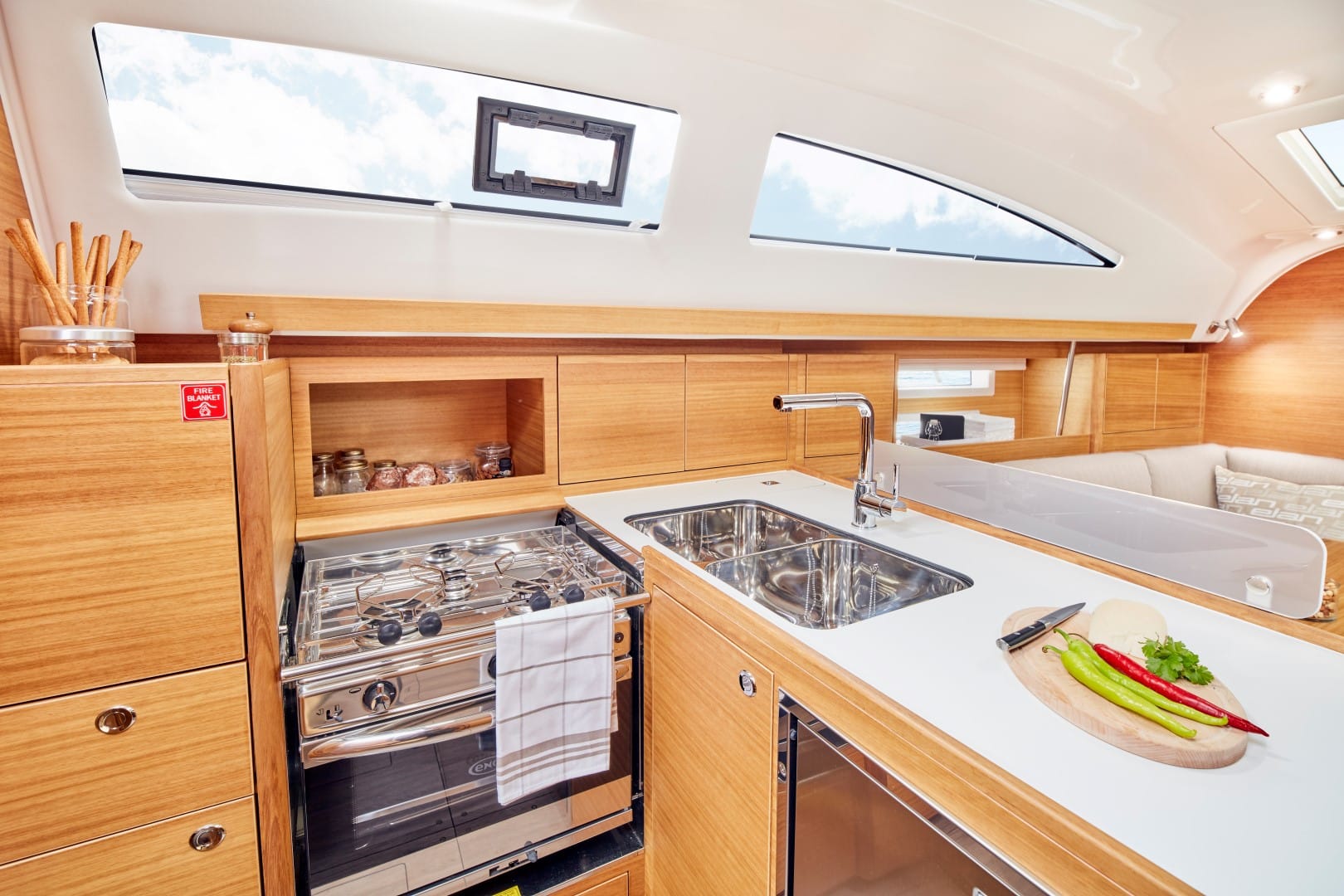 Innenraum einer modernen Segelyachtküche mit Holzschränken, einer Edelstahlspüle, einem Herd und frischem Gemüse auf einem Schneidebrett, mit natürlichem Licht, das durch große Fenster hereinströmt.
