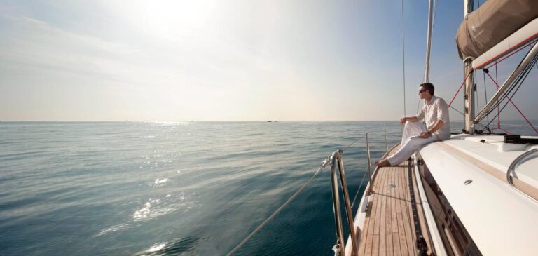 Ein Mann in weißer Kleidung entspannt sich auf dem Deck einer Segelyacht und blickt auf das ruhige Meer unter einem strahlenden Himmel. Die Sonne glitzert auf dem Wasser und unterstreicht die ruhige Atmosphäre.