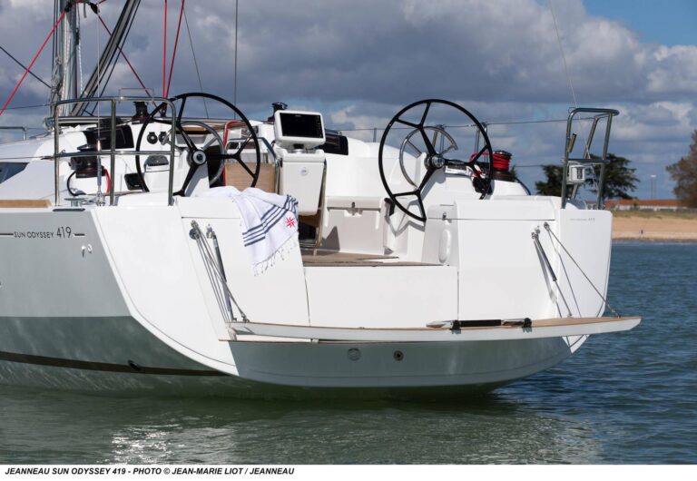 Rückansicht eines Segelboots Jeanneau Sun Odyssey 419, das in der Nähe eines Sandstrandes angedockt ist. Es verfügt über zwei Steuerräder und ein ordentlich organisiertes Deck, bereit für einen Segeltörn.