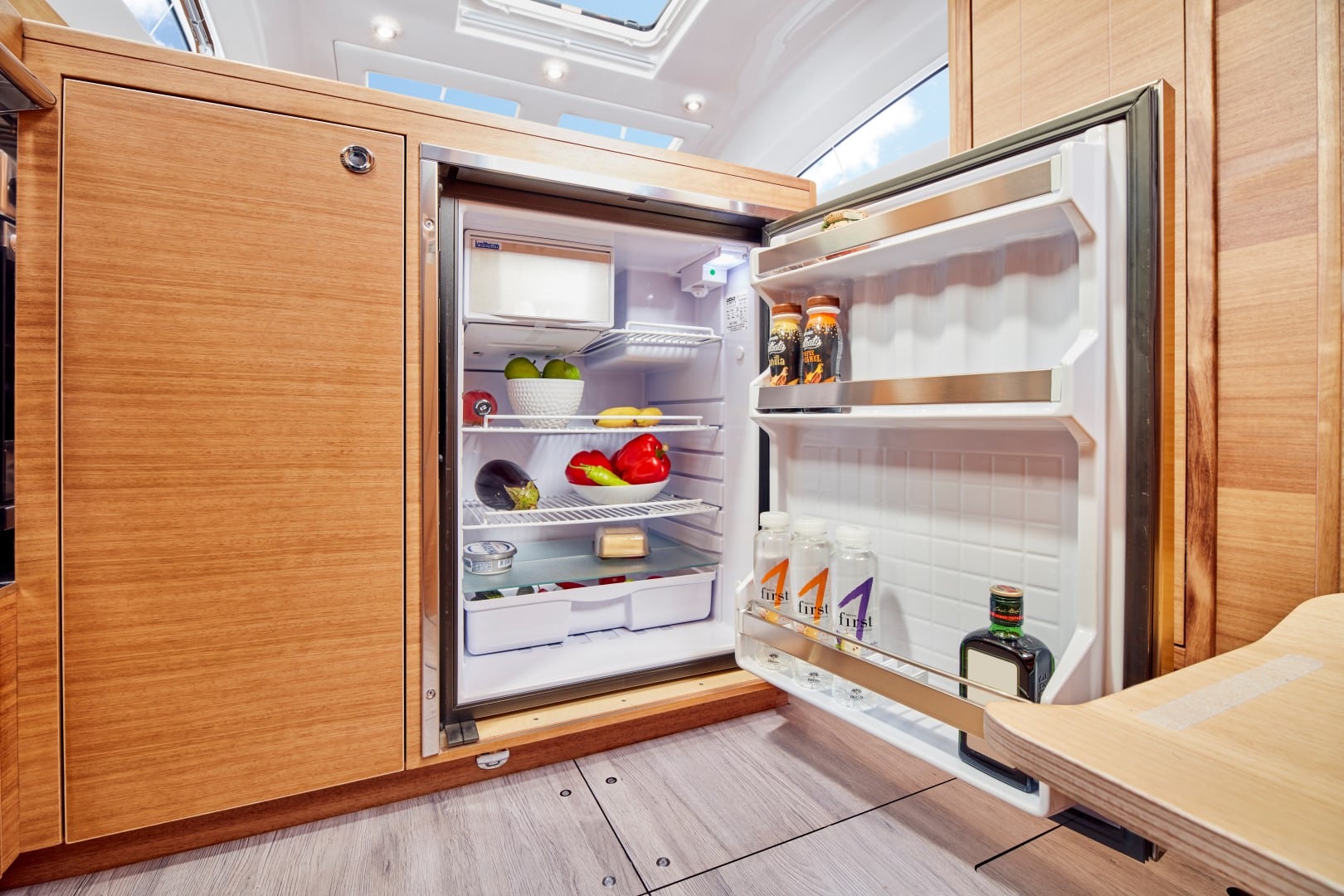Offener Kühlschrank gefüllt mit Lebensmitteln wie Obst und Getränken, eingebaut in einem Holzschrank im gut beleuchteten, modernen Innenraum einer Segelyacht.