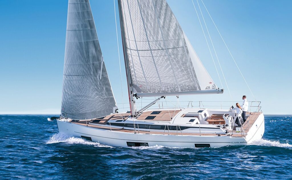 Eine moderne Segelyacht mit zwei Personen an Bord auf ruhiger See. Die Yacht hat große weiße Segel, die vollständig vor einem klaren blauen Himmel ausgerollt sind.