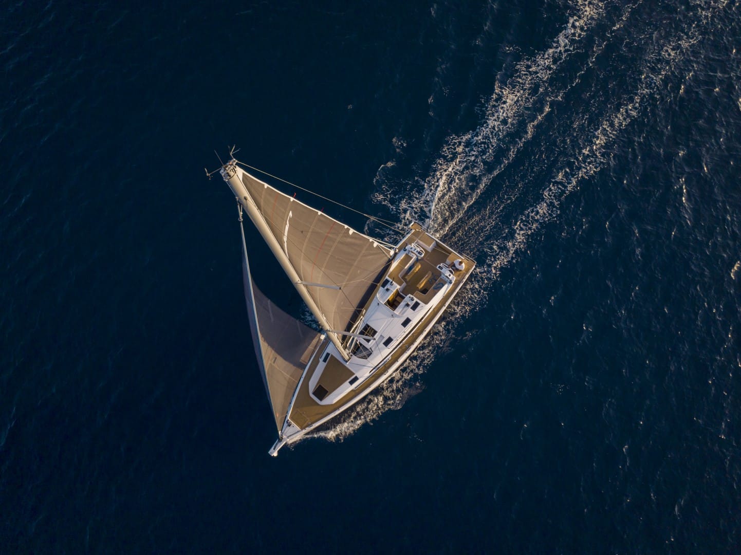 Luftaufnahme einer Segelyacht, die mit voll ausgefahrenen Segeln durch den Ozean schneidet und eine Heckwelle im klaren, tiefblauen Wasser erzeugt.