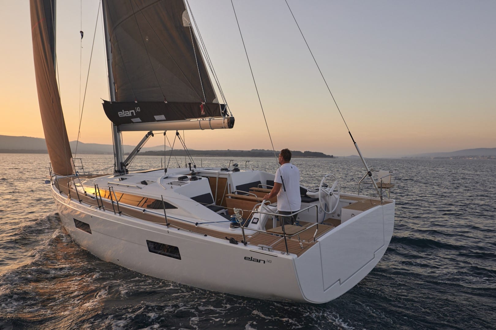 Ein Mann steuert eine moderne Segelyacht bei Sonnenuntergang, umgeben von ruhiger See und Hügeln am Horizont. Das Segelboot hat einen weißen Rumpf und ein schlankes Design.