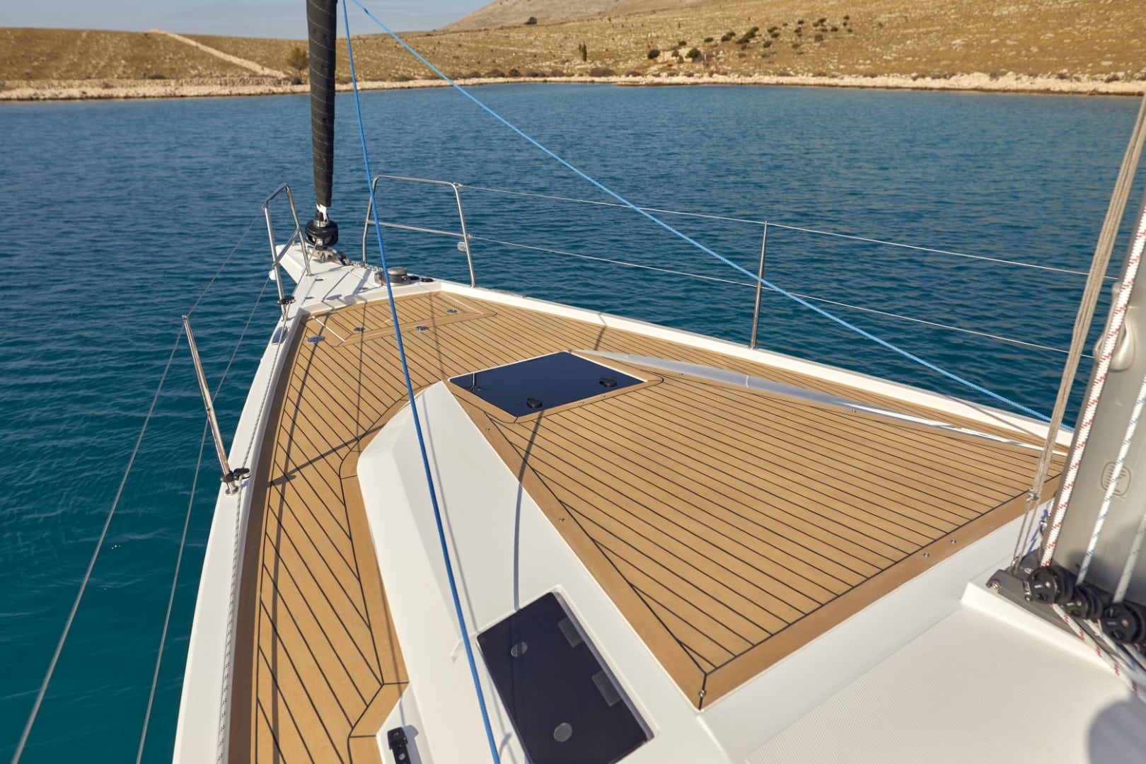 Der Bug einer Segelyacht blickt auf ein ruhiges blaues Meer, mit Holzdeckboden und ordentlich angeordneten Seilen, gegenüber einer kargen Küste unter einem klaren Himmel während eines Segeltörns.