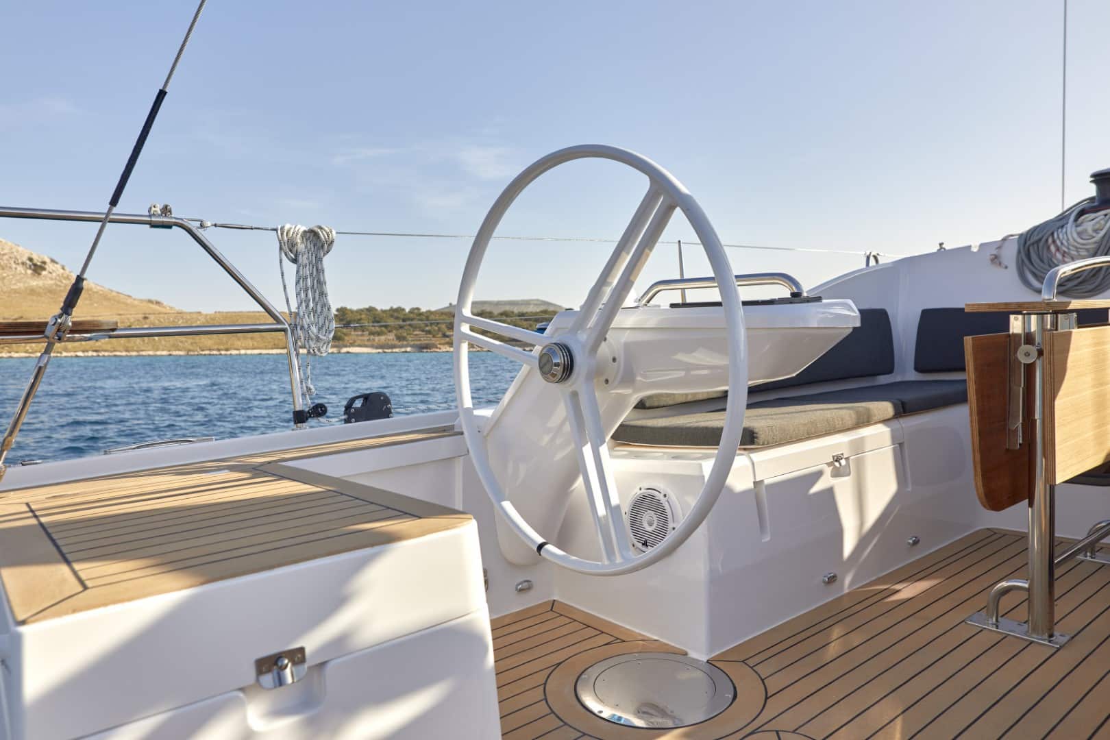 Das Steuerrad einer Yacht befindet sich an der Steuerbordseite und bietet Aussicht auf das ruhige Meer und die Küste. Das Deck ist mit poliertem Holz verkleidet und Segeltaue sind ordentlich in der Nähe aufgerollt, bereit für einen Segeltörn.