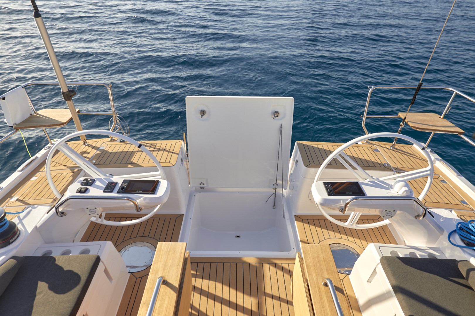 Hinteres Deck eines Katamarans mit zwei Steuerrädern und Navigationsbildschirmen, mit Blick auf ruhiges blaues Wasser. Das Deck verfügt über Holzboden und eine weiße Sitzbank.