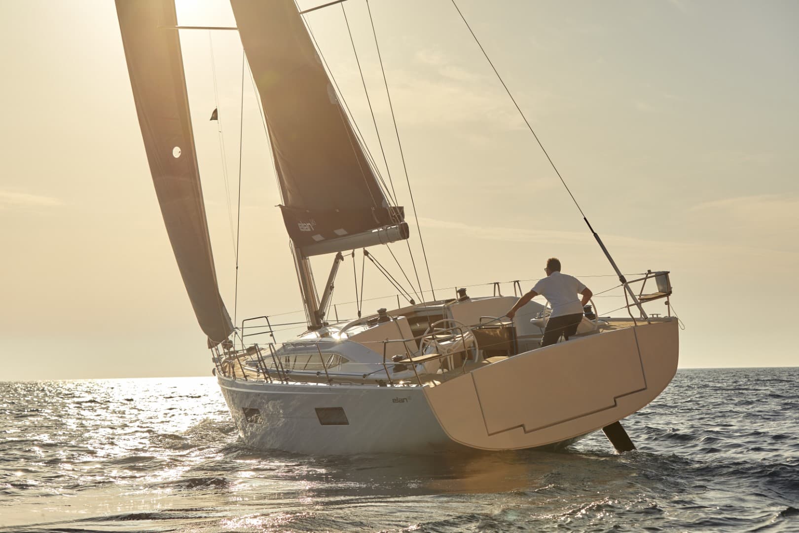 Eine Person steht am Heck einer Segelyacht und steuert das Boot, während es vor der Kulisse eines Sonnenuntergangs anmutig auf ruhiger See segelt.