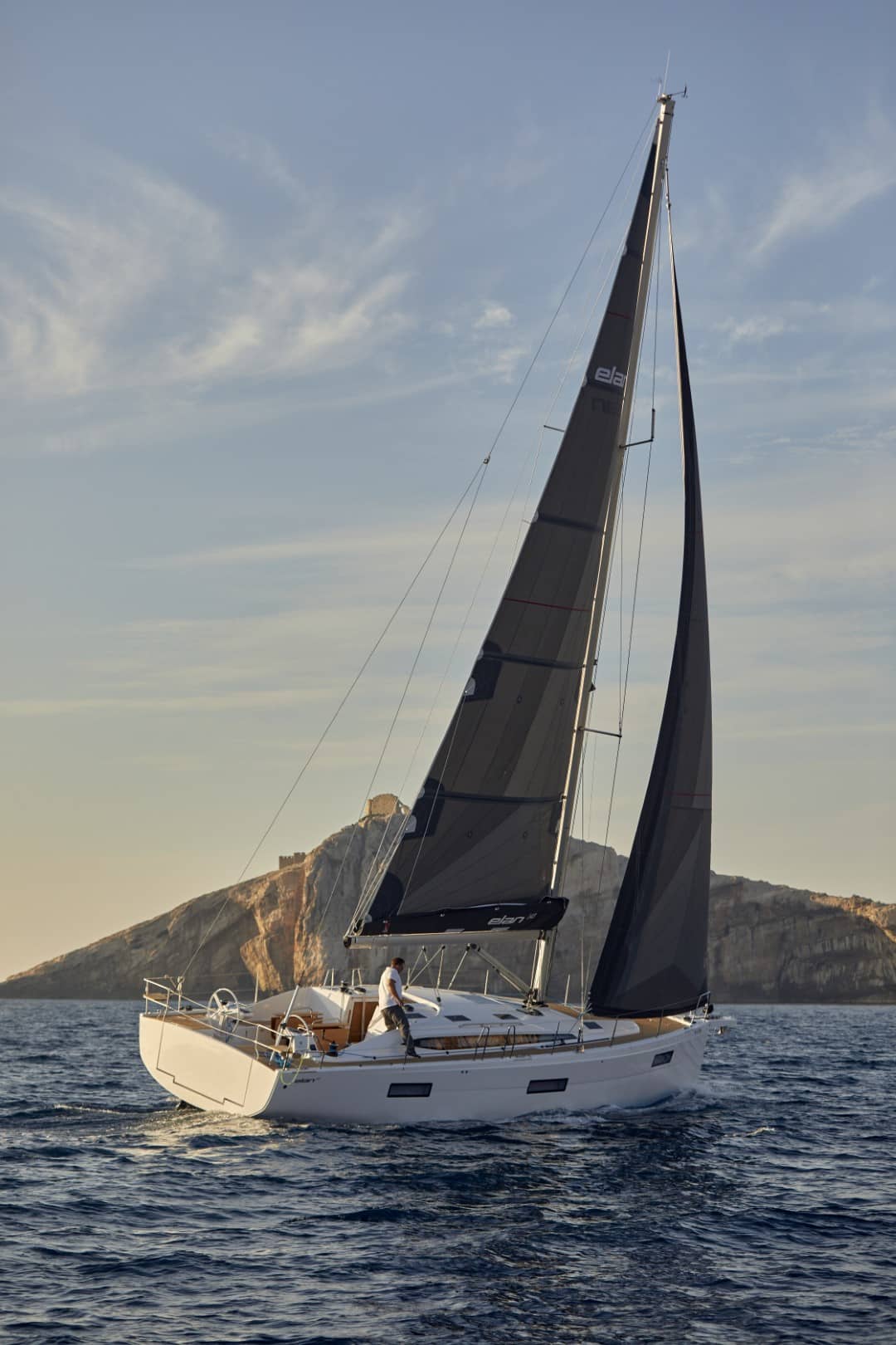 Eine Segelyacht mit weißem Rumpf und dunklen Segeln gleitet während der goldenen Stunde nahe einer felsigen Küste durch das Wasser. Darüber ist ein klarer Himmel und an Deck ist eine Person zu sehen.