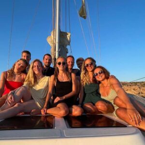 Eine Gruppe von neun Personen, die zusammen lächeln und auf einer Segelyacht unter einem klaren blauen Himmel posieren. Sie sitzen auf dem Deck, im Hintergrund sind felsige Landschaft und Wasser zu sehen. Alle wirken entspannt und glücklich und genießen ihren Segeltörn im Freien.