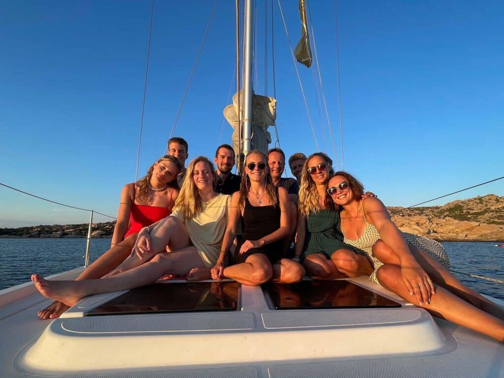 Eine Gruppe von acht Personen, fünf Frauen und drei Männer, sitzt zusammen auf dem Deck eines Katamarans unter einem klaren, sonnigen Himmel. Sie lächeln und posieren für das Foto, mit einer malerischen Kulisse aus ruhigem Wasser und felsigem Gelände in der Ferne während ihres Segelurlaubs.