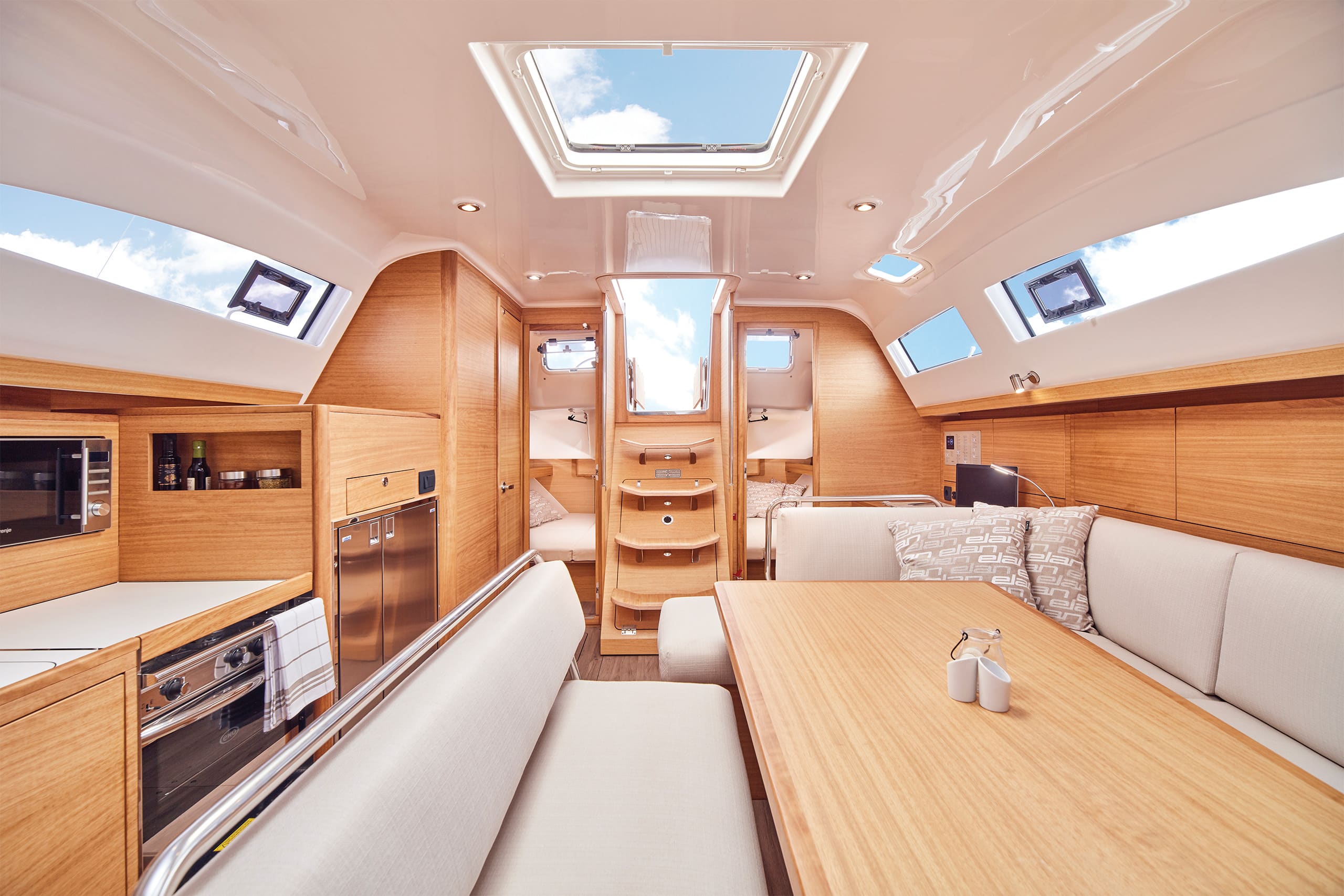 Das Bild zeigt das Innere einer modernen Segelyachtkabine. Es verfügt über helle Holzmöbel, einen Essbereich mit Tisch und gepolsterten Sitzbänken, eine Küchenzeile mit Geräten und eine Treppe, die zum Deck führt. Natürliches Licht erhellt den Raum durch Oberlichter und lädt die Gäste zum gemütlichen Mitsegeln ein.