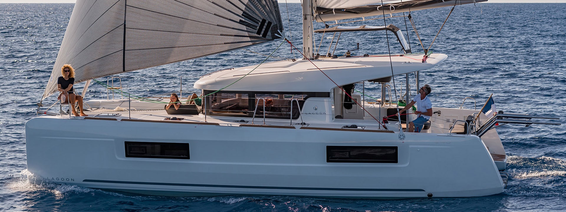 Ein moderner Katamaran segelt auf einem ruhigen, blauen Meer. Mehrere Personen entspannen sich an Deck und genießen den Segeltörn unter sonnigem Himmel. Eine Frau steht vorne.