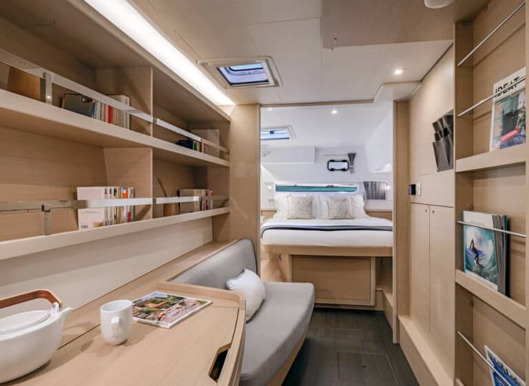 Innenansicht einer modernen Segelyachtkabine mit einem gemütlichen Schlafzimmer mit großem Bett, einem Wohnbereich mit grauem Sofa und Regalen voller Bücher und Dekoartikel. Helle Beleuchtung und Holzpaneele werten den Raum auf.