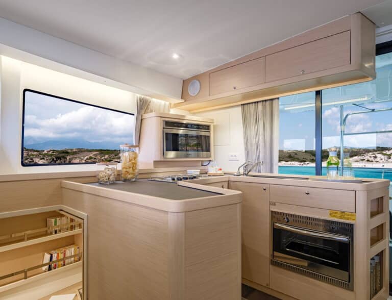 Moderne Bootsküche mit hellen Holzschränken, eingebauter Mikrowelle, Backofen und einer Theke. Ein großes Fenster gibt den Blick auf eine sonnige Landschaft während eines Segeltörns frei. Weingläser und eine offene Snackdose stehen auf der Theke.