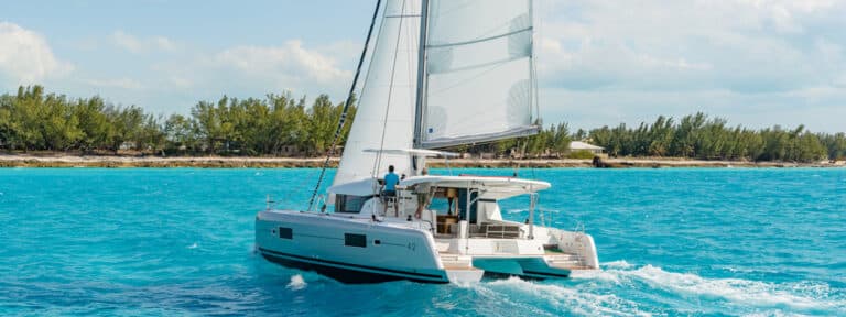 Eine Segelyacht mit weißen Segeln kreuzt in der Nähe einer tropischen Insel mit klarem blauen Wasser und einigen sichtbaren Bäumen an der Küste unter einem teilweise bewölkten Himmel.