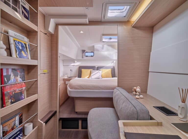 Innenansicht einer modernen Katamarankabine mit gemütlichem Doppelbett, stilvoller Holzvertäfelung, großen Regalen mit Büchern und einem grauen Sofa. Natürliches Licht fällt durch die Luken oben ein.