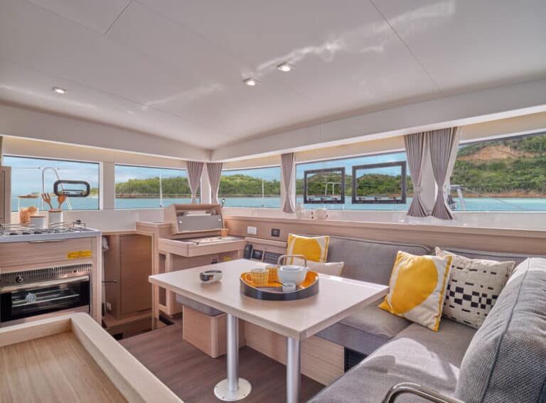 Innenansicht einer modernen Yacht mit einer Küchenzeile mit Holzschränken und Geräten aus Edelstahl, einem Essbereich mit einem grauen Sofa, gelben Kissen und einem weißen Tisch, alles mit Blick auf das Meer durch große Fenster. Ideal für einen Segelurlaub oder ein Mitsegelabenteuer auf diesem eleganten Katamaran.