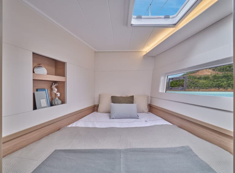 Ein gemütliches Kabinenschlafzimmer auf einem Katamaran mit einem kleinen Bett, weichen Kissen und einer Decke. Das Zimmer verfügt über ein Fenster mit Blick auf das Meer und ein kleines Regal mit Dekorationsgegenständen. Die Decke hat ein Oberlicht, das den Raum mit natürlichem Licht erhellt – perfekt für Ihren Segelurlaub.