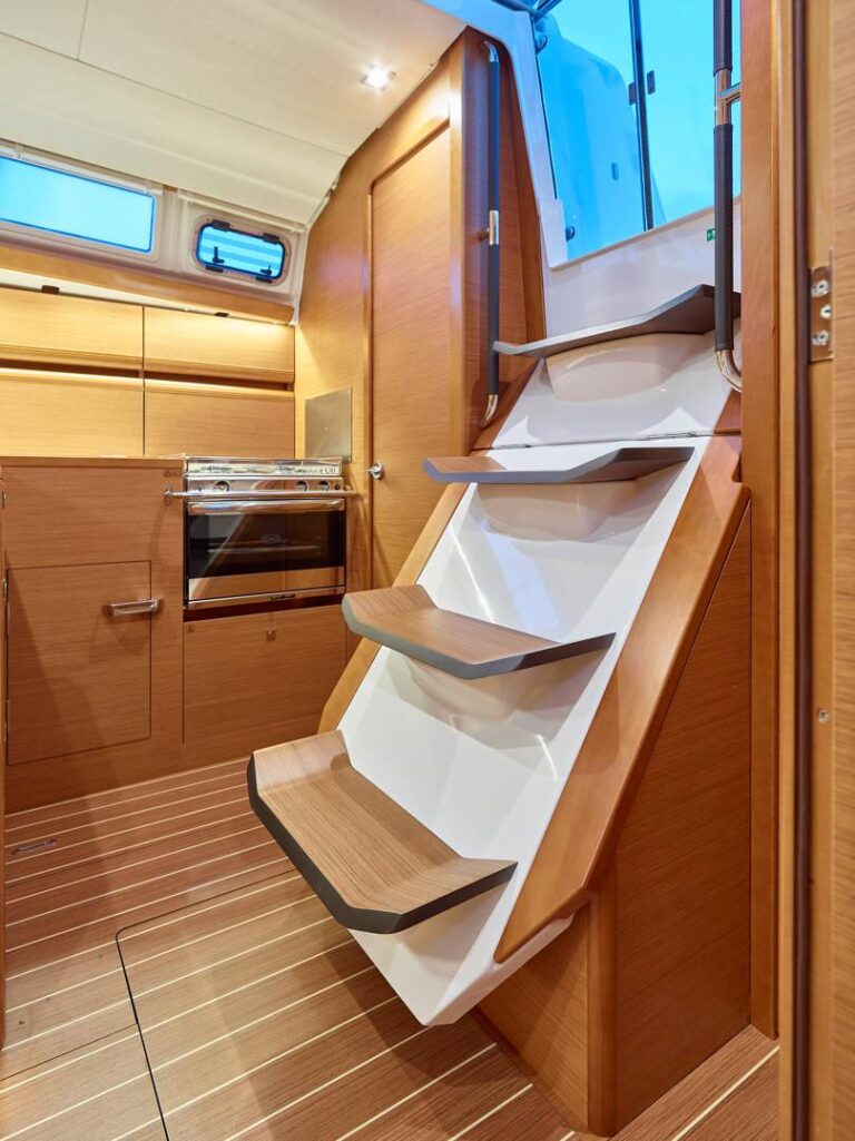 Innenansicht einer modernen Bootskabine mit Holzverkleidung. Das Bild zeigt eine schmale Treppe nach oben, daneben befindet sich ein kleiner Küchenbereich mit Herd, Schränken und Schubladen. Der Raum ist ideal für Ihren nächsten Segeltörn, ist mit Deckenbeleuchtung gut beleuchtet und strahlt sowohl Komfort als auch Stil aus.