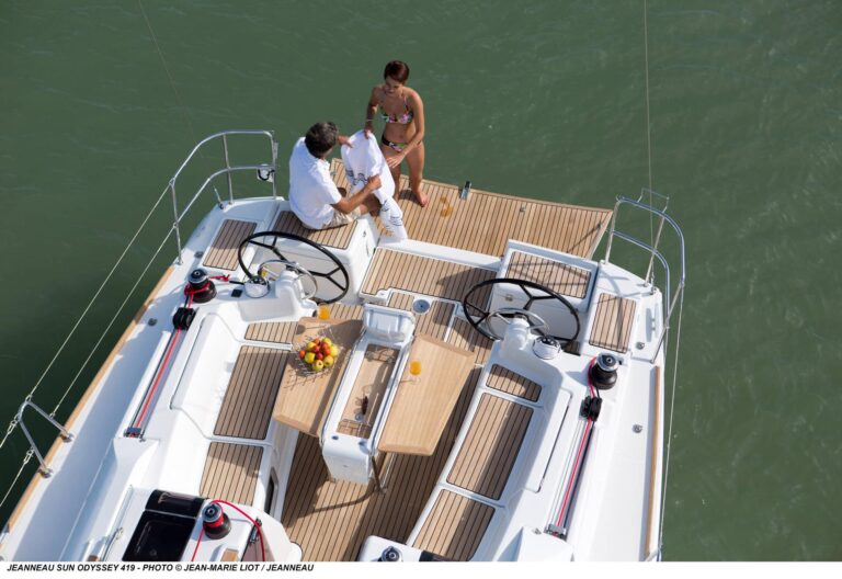 Zwei Personen genießen einen sonnigen Tag auf einer Luxus-Segelyacht. Der Mann steht am Steuer und die Frau sitzt daneben inmitten eines geräumigen Teakdecks. Wasser und Horizont sind sichtbar.