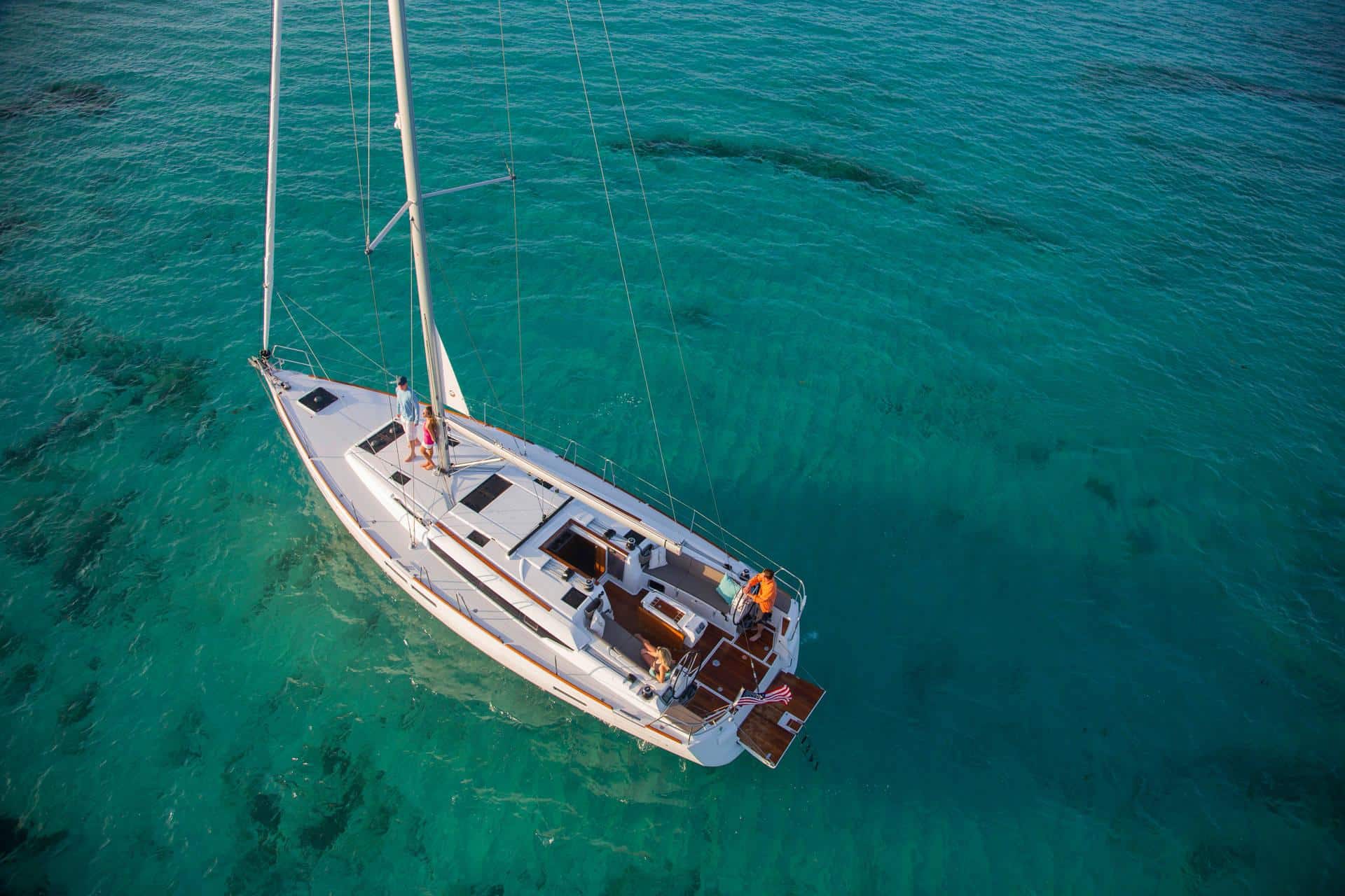 Luftaufnahme eines Segelboots mit weißen Segeln, das über klares, türkisfarbenes Wasser gleitet. An Bord der Segelyacht befinden sich zwei Personen, die den sonnigen Tag genießen. Das ruhige Meer darunter gibt dank der Klarheit des Wassers den Blick auf den Meeresboden frei und macht diese Segelreise zu einem perfekten Ausflug. Eindeutig mitsegeln exklusiv mit der maßgeschneiderten Segelreise.